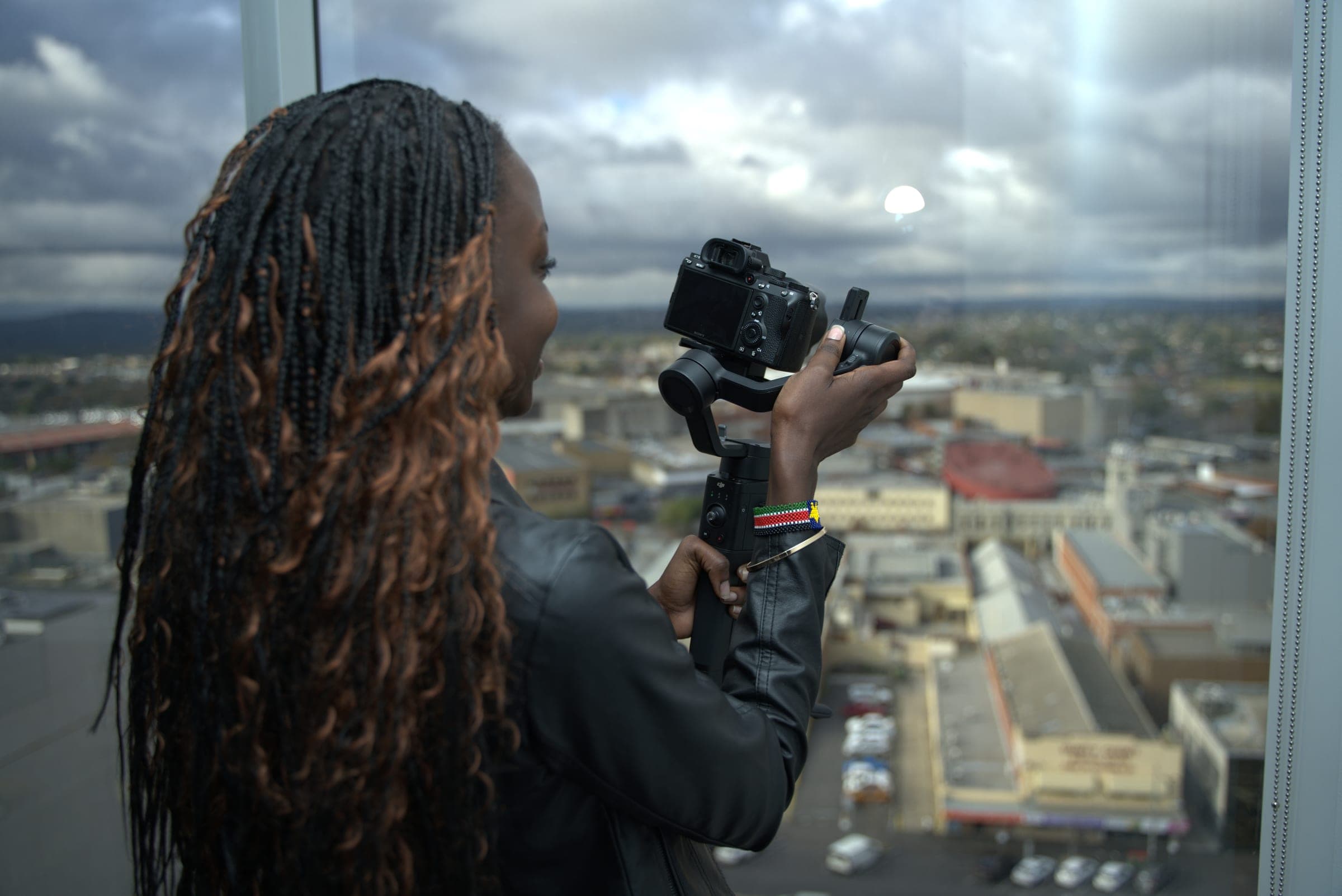 Akoi Chol, seen from behind, filming the city skyline from a high window with her camera on a gimbal.