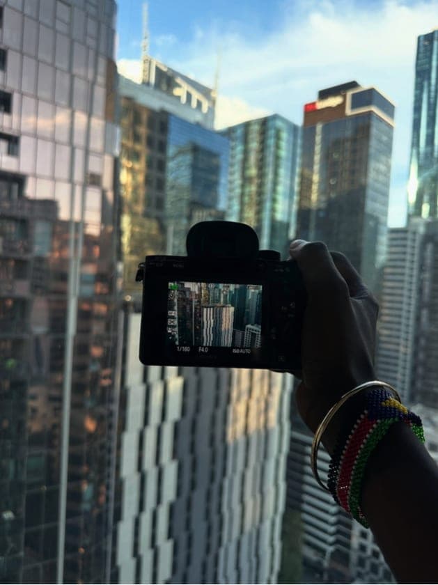 A hand with beaded bracelets holding up a camera whose screen frames the city towers.