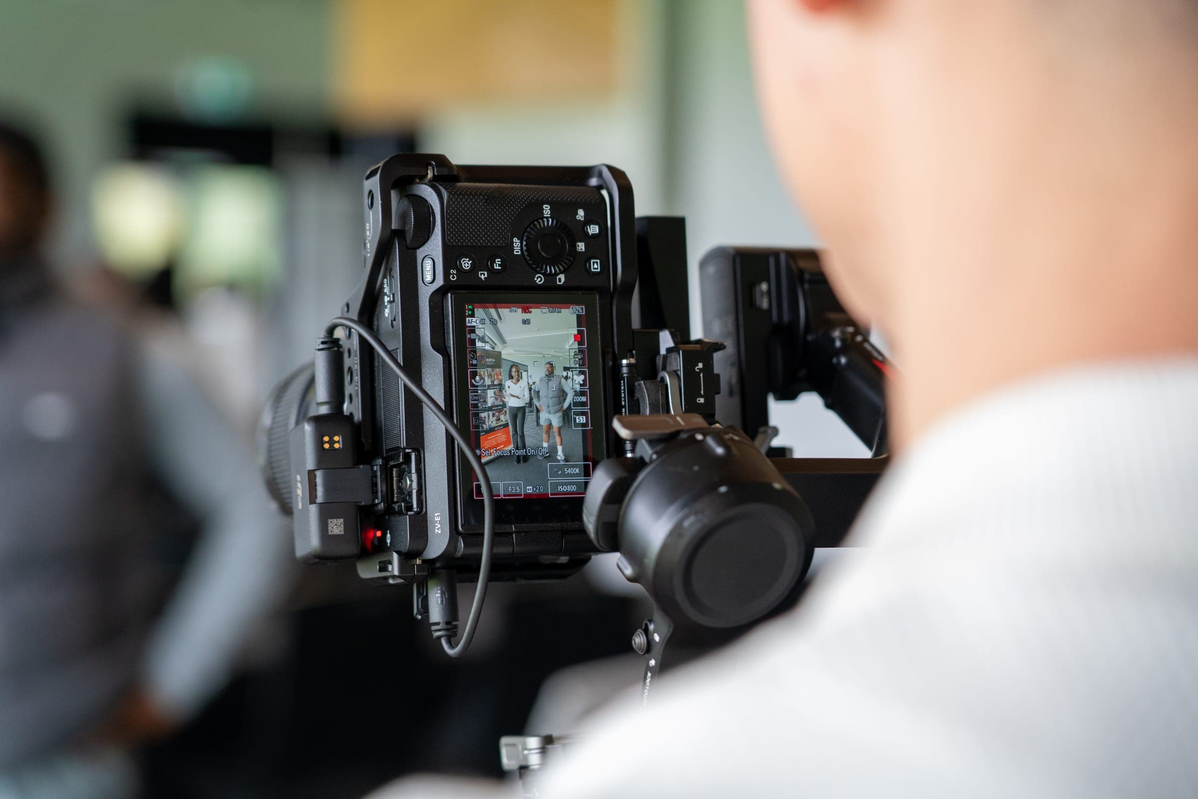 Over a shoulder, a camera on a gimbal, its screen showing two people being filmed.