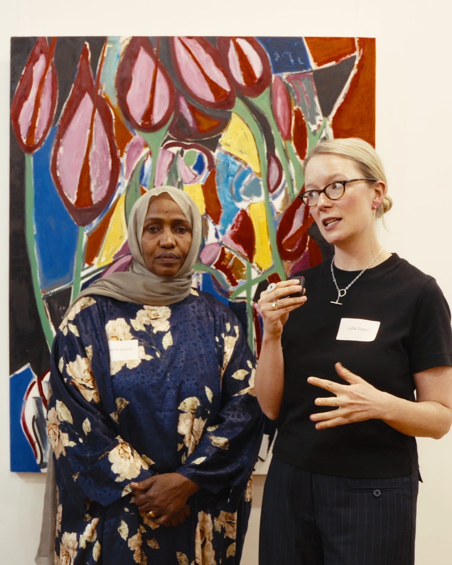 At Inner North Community Foundation's event, a woman in black speaks in front of a bold painting of pink tulips, beside a woman in a headscarf and a floral dress who listens.