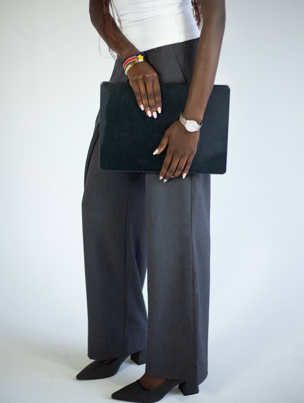 In the studio, a woman in grey wide-leg trousers and a white top holds a black laptop sleeve at her hip.