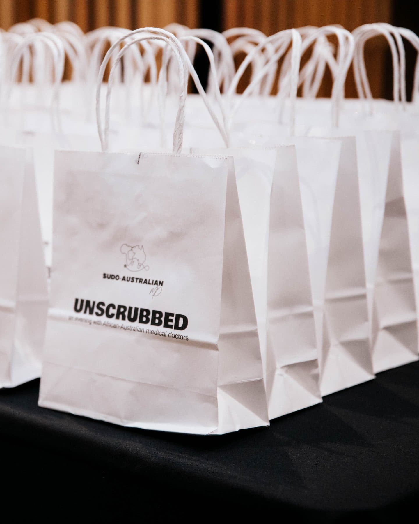 White gift bags lined up on a black table, printed “UNSCRUBBED: an evening with African-Australian medical doctors”.