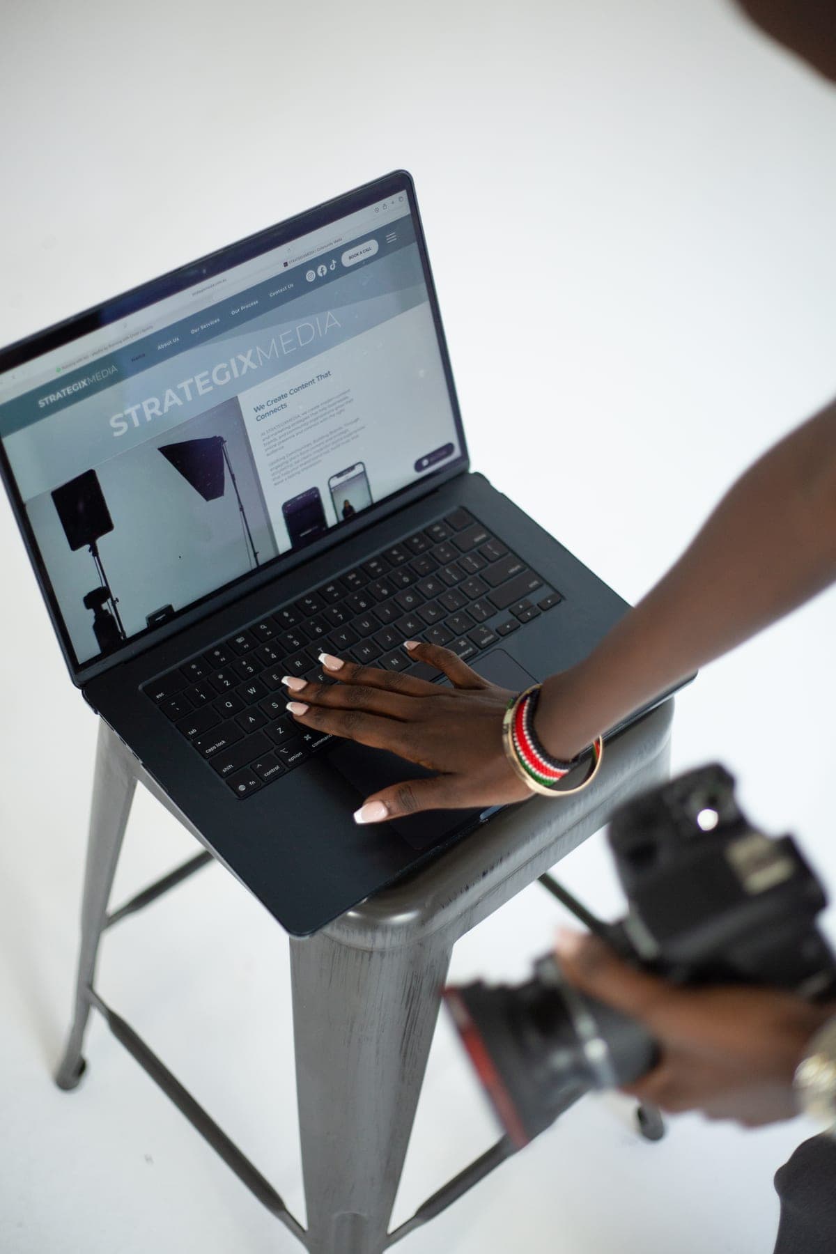 A hand on a laptop resting on a studio stool, with a camera held close by.
