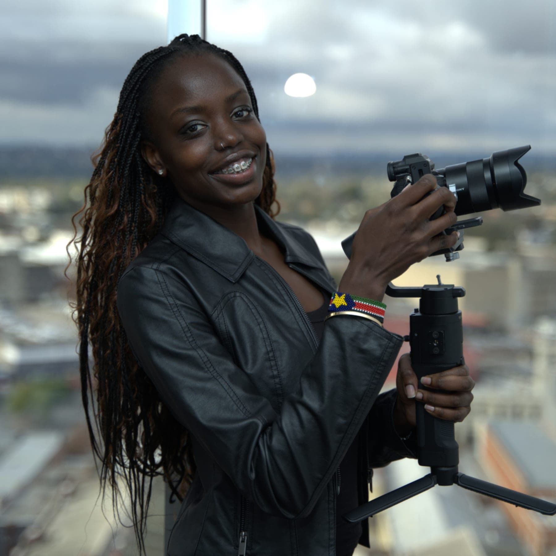 Akoi Chol smiling, holding her camera on a gimbal, with the city behind her.