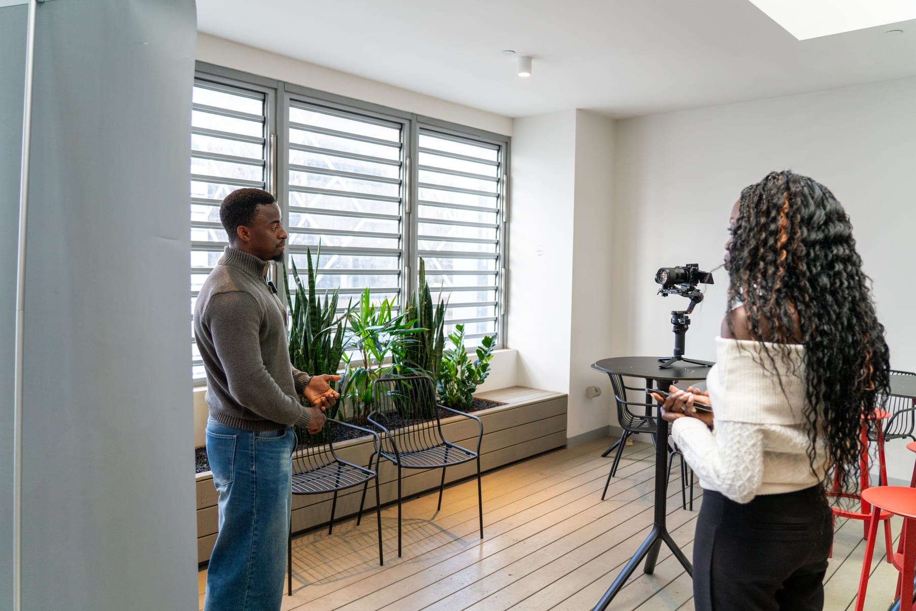 Akoi Chol on a video shoot in a bright office, her camera on a gimbal, as a man in a grey jumper talks with her.