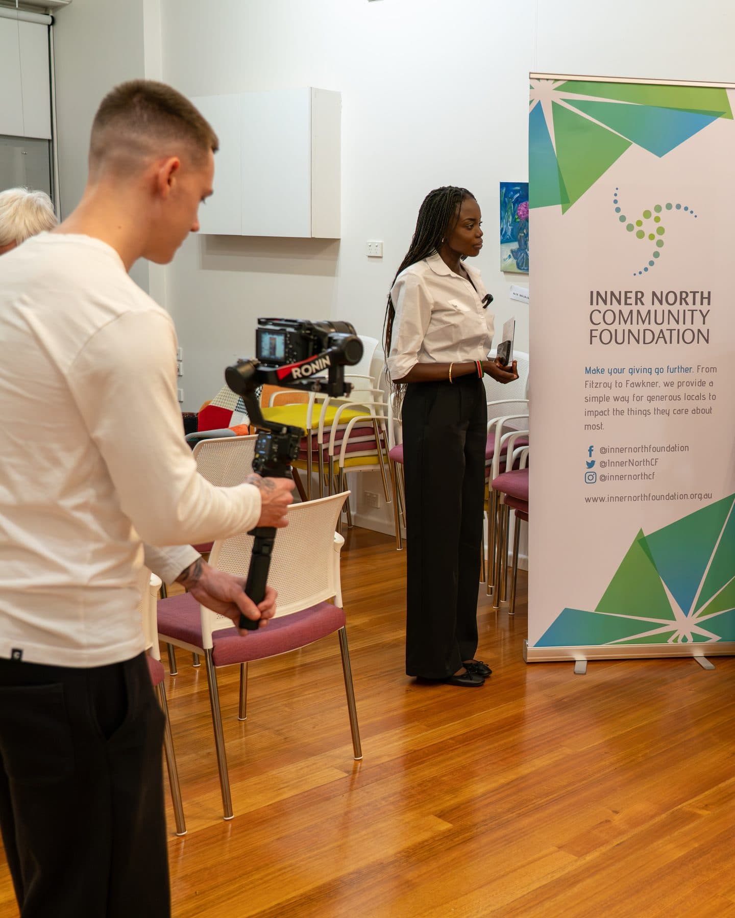 Behind the scenes at Inner North Community Foundation's event: a videographer filming with a camera on a gimbal, and Akoi Chol beside the foundation's banner.