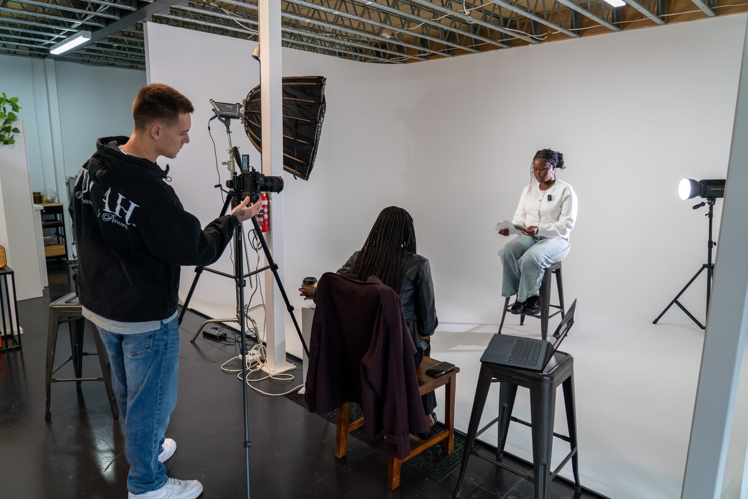 A videographer films a woman reading from notes on a stool in a white studio, as someone with long braids watches from a chair.